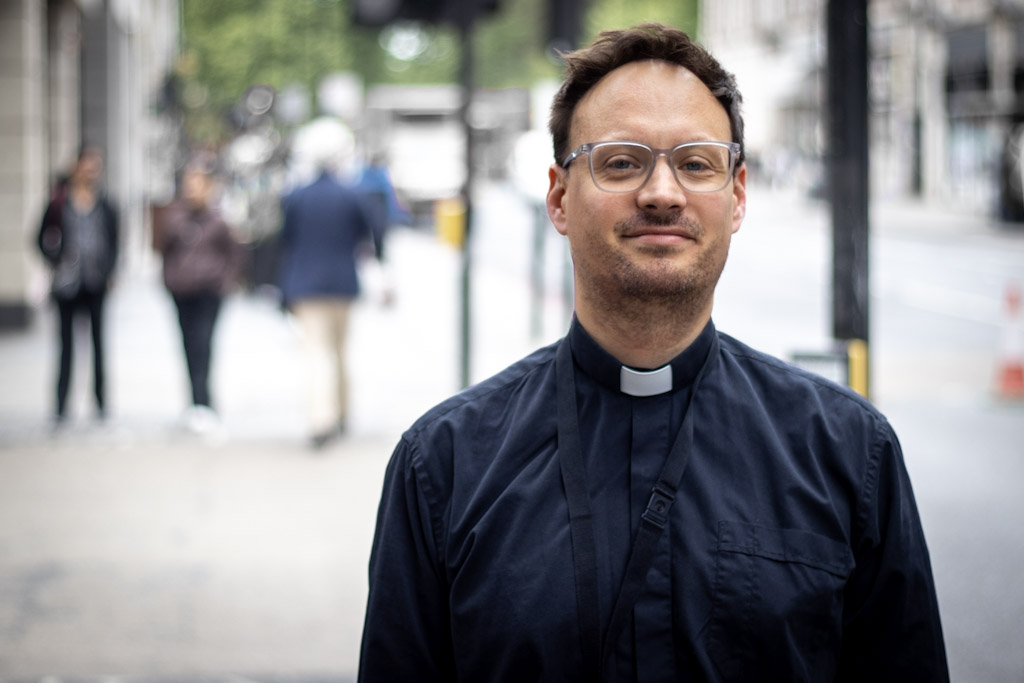 A man wearing glasses and a clergy collar stands in the foreground, with people walking in the background on a city street.
